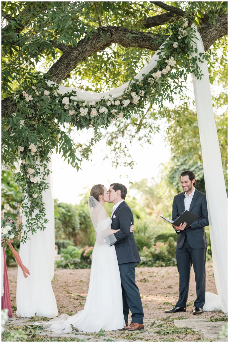 Barr Mansion Wedding Photographer captures first kiss as husband and wife at outdoor ceremony in Austin Texas with floral garland and fabric draping the tree behind them