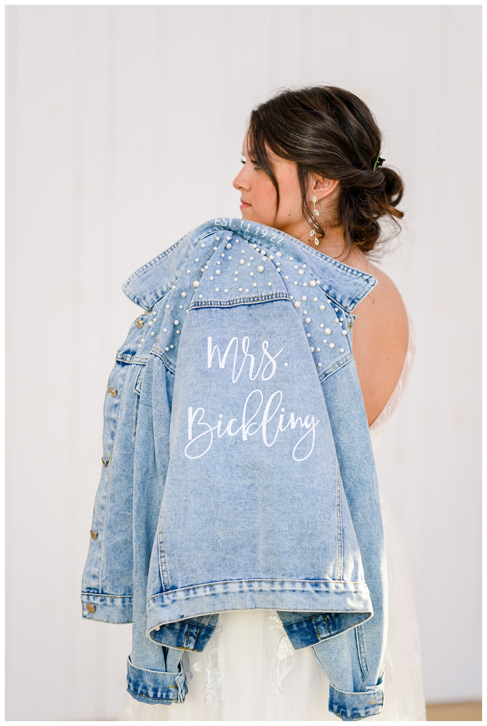 Bride holds denim jacket with new last name embroidery on the back with pearls over her shoulder during bridal portraits at wish well house wedding venue in Georgetown Texas