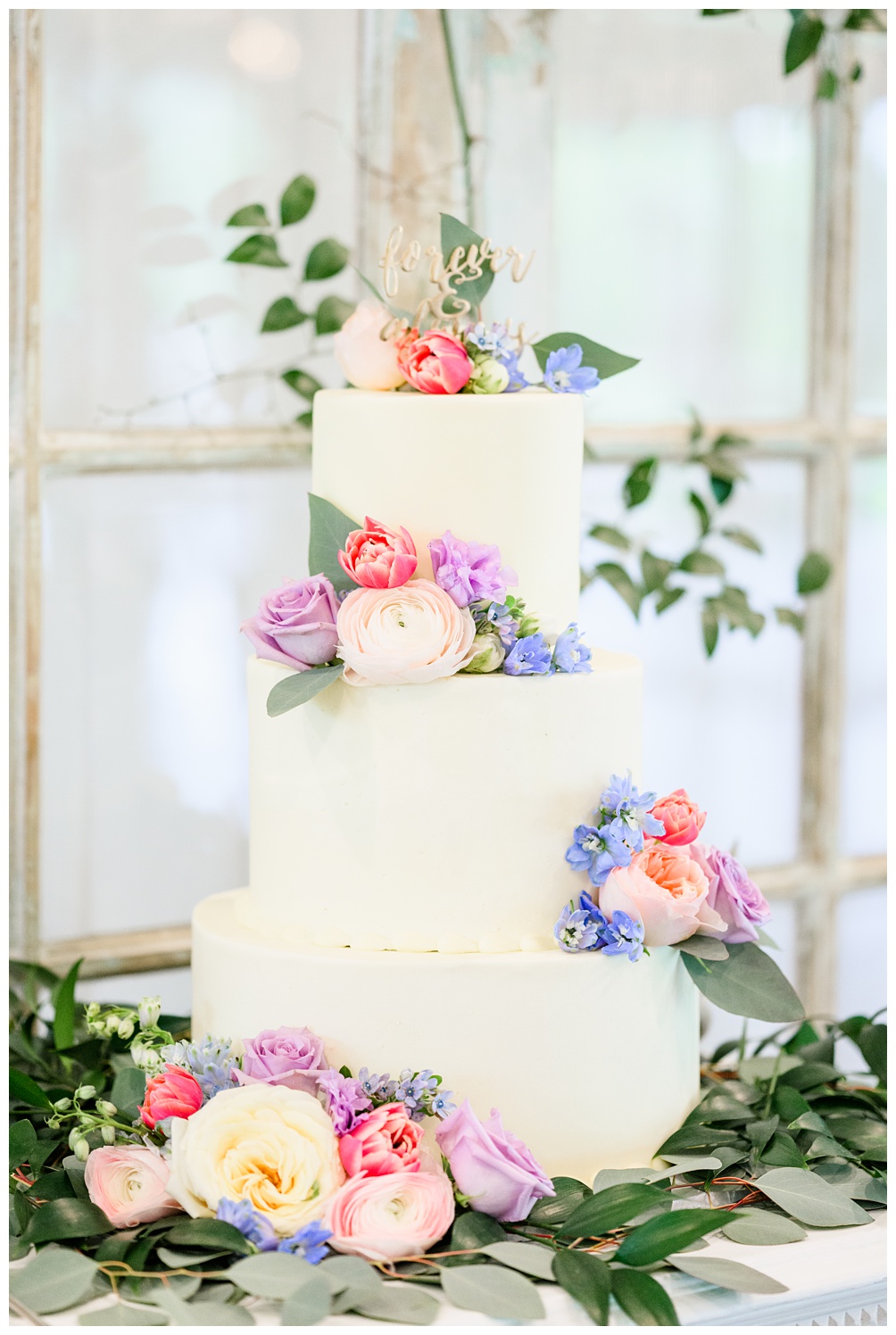 The Hayley Cakes wedding cake covered in Spring flowers and topped with gold sign that says forever and always