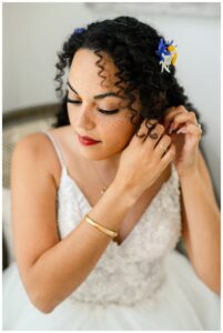 Close-up of bride putting on earrings, wearing a lace wedding dress and soft floral hair accessories, with curly hair and natural glam makeup during her Austin bridal portraits at the Gruene Estate with Mylah Renae.