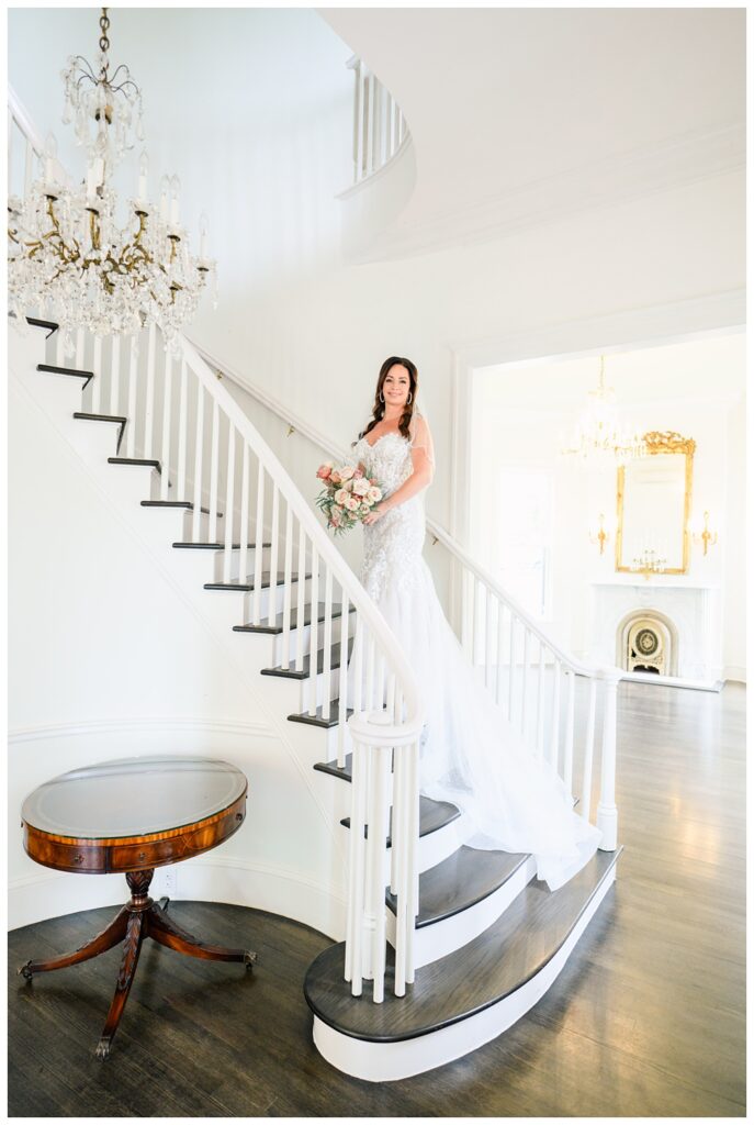Bride stands on a sweeping staircase beneath a crystal chandelier, wearing a fitted lace wedding dress with a long train and holding a blush bouquet during her elegant Austin bridal portrait session.