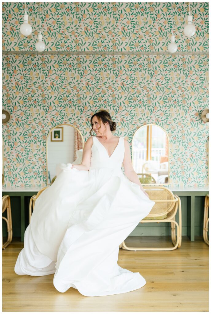 Bride twirls in a voluminous, modern wedding gown with a deep V neckline, framed by colorful patterned wallpaper and rattan chairs during a joyful Austin bridal shoot at Wish Well House.