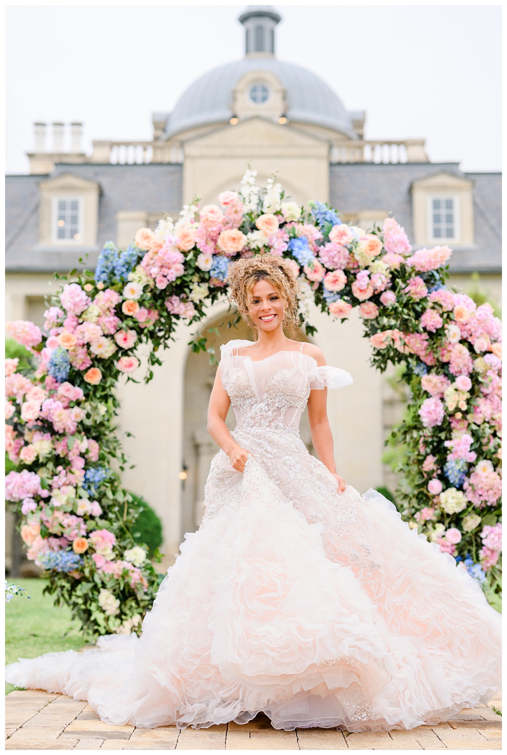 Bride poses in a voluminous blush and ivory wedding gown with off-the-shoulder sleeves, standing beneath a whimsical pink and blue floral arch at a European-style Austin estate.