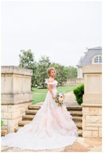 Bride in a dramatic ruffled train walks down stone steps holding a pastel bridal bouquet, with the romantic architecture of an Austin estate behind her.