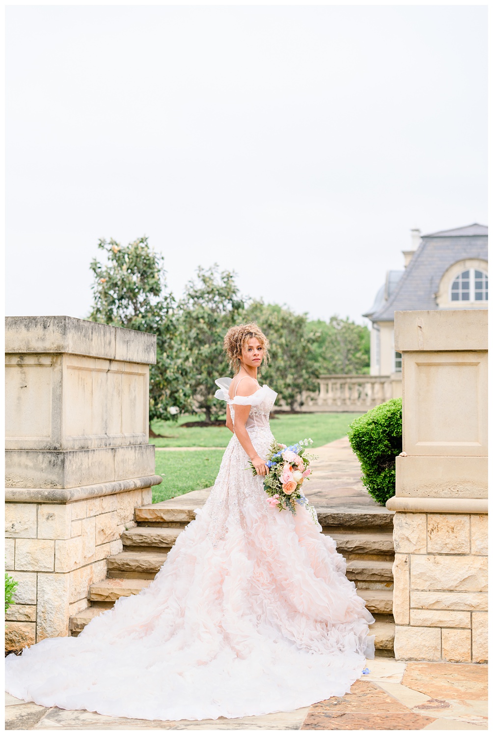 Bride in a dramatic ruffled train walks down stone steps holding a pastel bridal bouquet, with the romantic architecture of an Austin estate behind her.