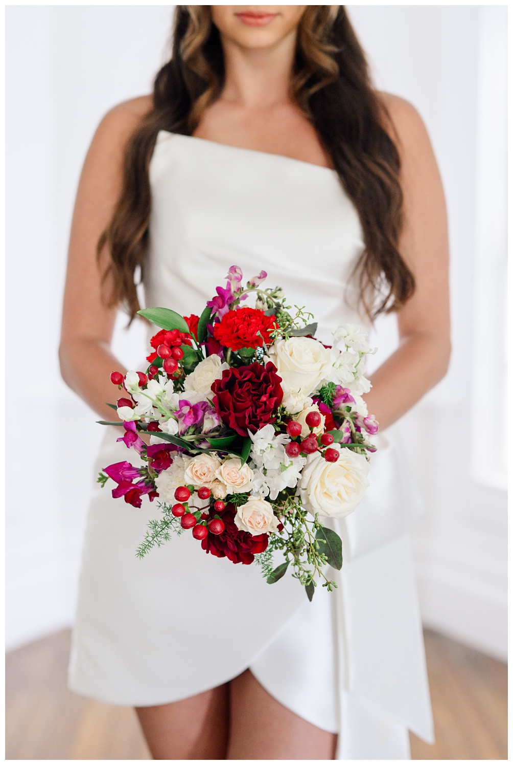 Close-up of a modern bride in a white one-shoulder mini dress holding a bold bouquet with red ranunculus, ivory roses, berries, and playful pink florals during an Austin bridal shoot.