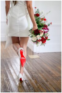 Bride walks across a wooden floor wearing a white bridal mini dress and Christian Louboutin heels, holding a bold red and ivory bouquet during a modern Austin bridal portrait session.