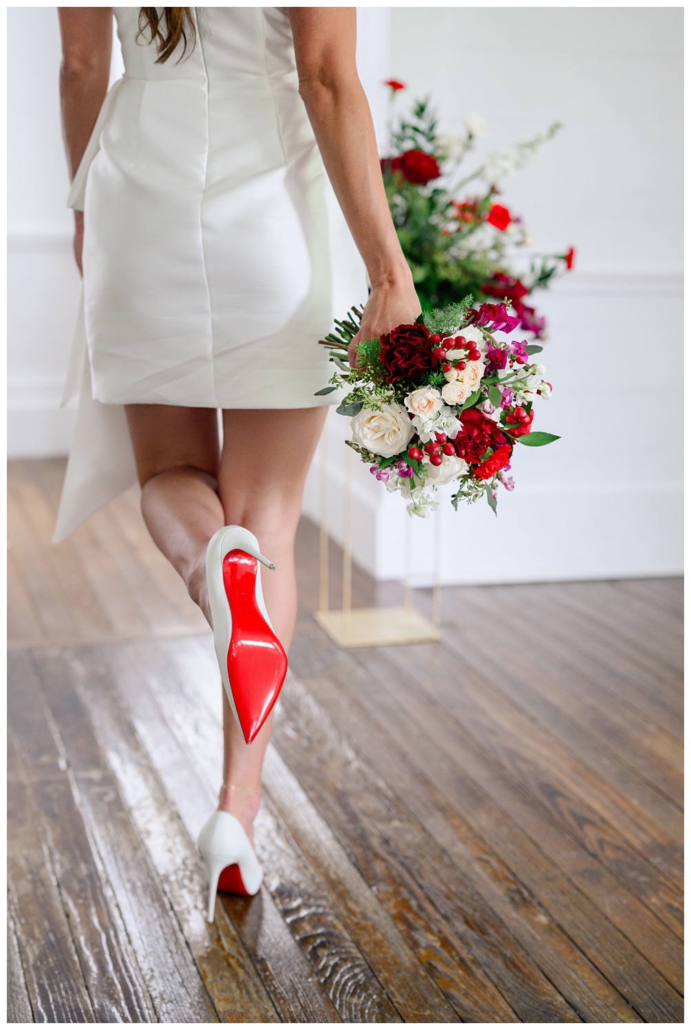 Bride walks across a wooden floor wearing a white bridal mini dress and Christian Louboutin heels, holding a bold red and ivory bouquet during a modern Austin bridal portrait session.