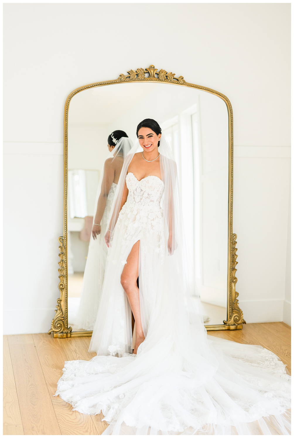 Bride poses confidently in front of an ornate gold mirror in a strapless lace gown with a thigh-high slit and sheer veil during her Austin bridal photoshoot.