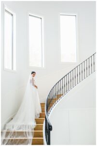 Bride stands on a sweeping staircase in a timeless wedding gown with a full train, captured during her elegant indoor Austin bridal session.