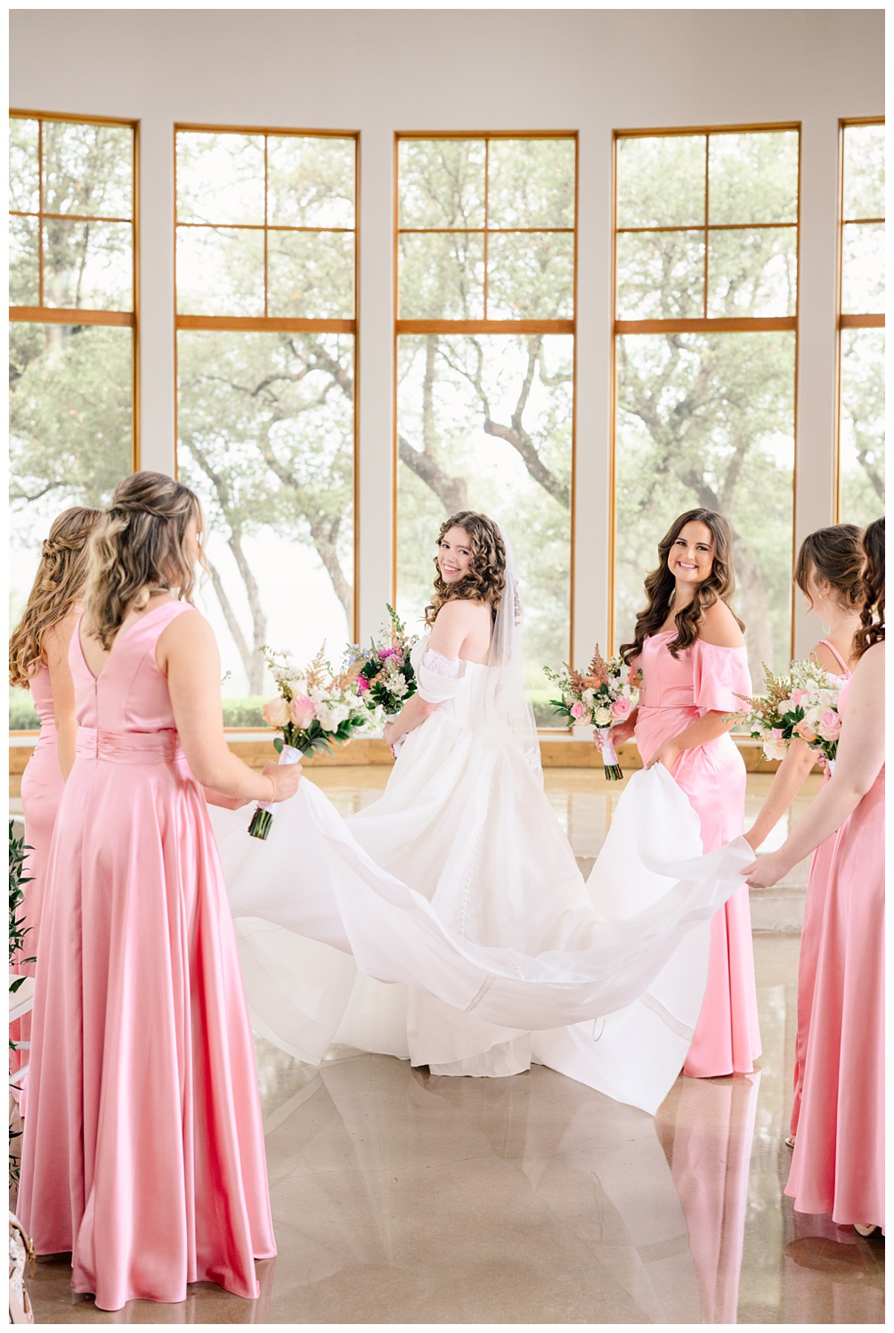 Bridesmaids in pink dresses help bride by carrying her dress to ceremony site of Canyonwood Ridge Wedding